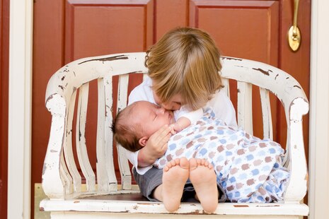 Zum fachlichen Einstieg bzw. als Vorbild zur Rubrik Kindergeld ist stellvertretend ein Bild dargestellt. Auf dem Bild ist ein kleineres Kind auf einer Bank sitzend abgebildet, welches in seinem Arm ein Baby hält. Diesem drückt er einen Kuss auf die Wange.